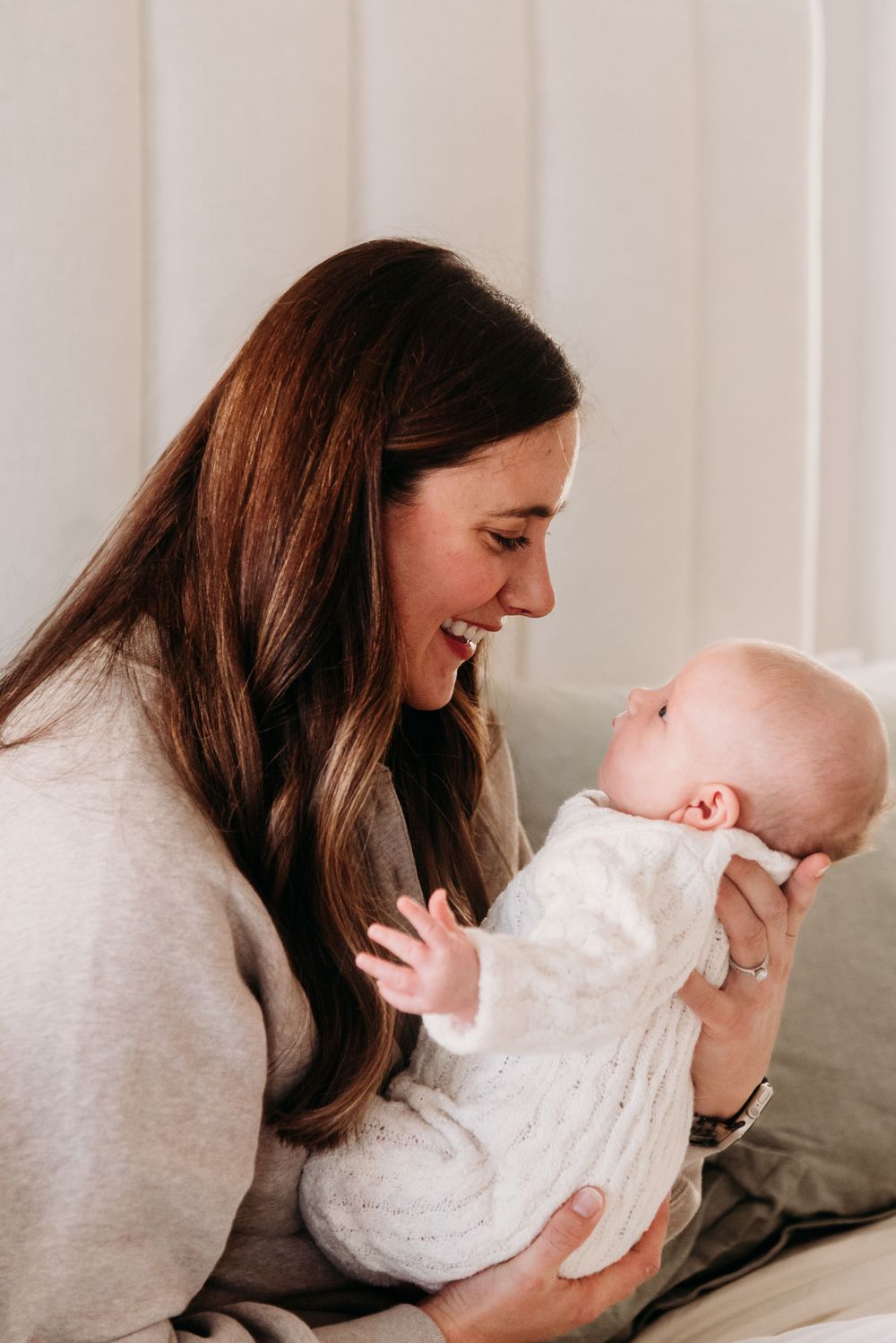 A person in a grey sweater smiles while holding a baby wearing white clothing in a bright indoor setting.