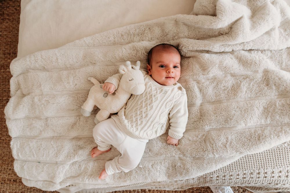 A baby lies on a white knitted blanket wearing a cozy white cable knit sweater with a stuffed animal companion.