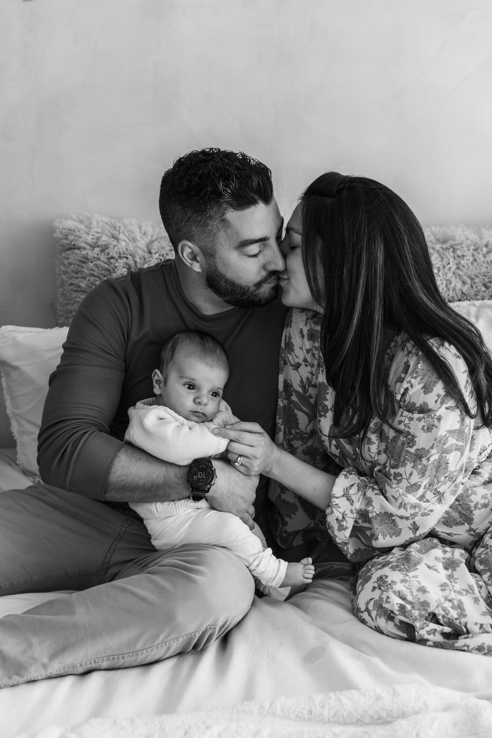 Black and white photo of a couple sharing an intimate moment on a bed with their infant baby in a tender family scene.