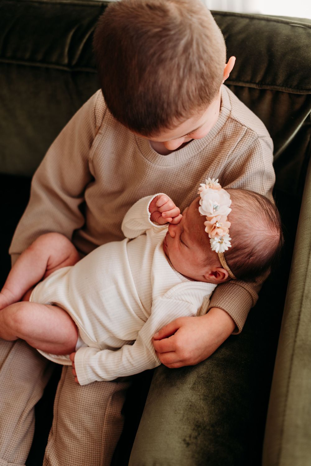 A young child holds and gazes down at a newborn baby wearing a white floral headband on a dark green couch.
