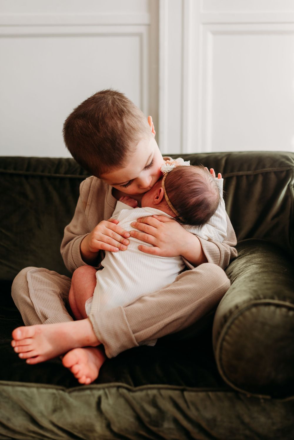 A young child gently embraces and kisses a newborn baby while sitting on a dark green couch.