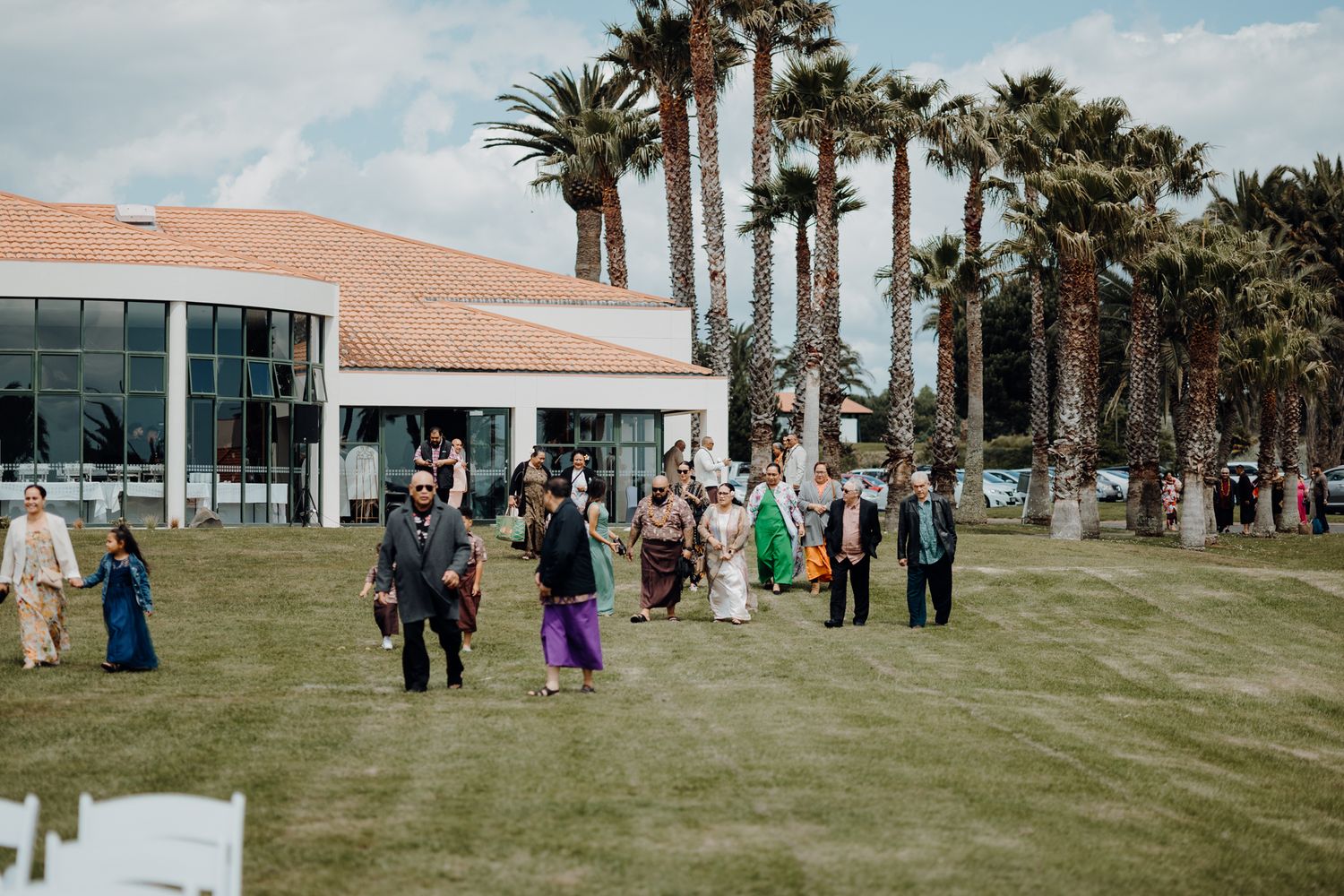 Wedding guests gather on a lush green lawn with palm trees outside a modern venue building.