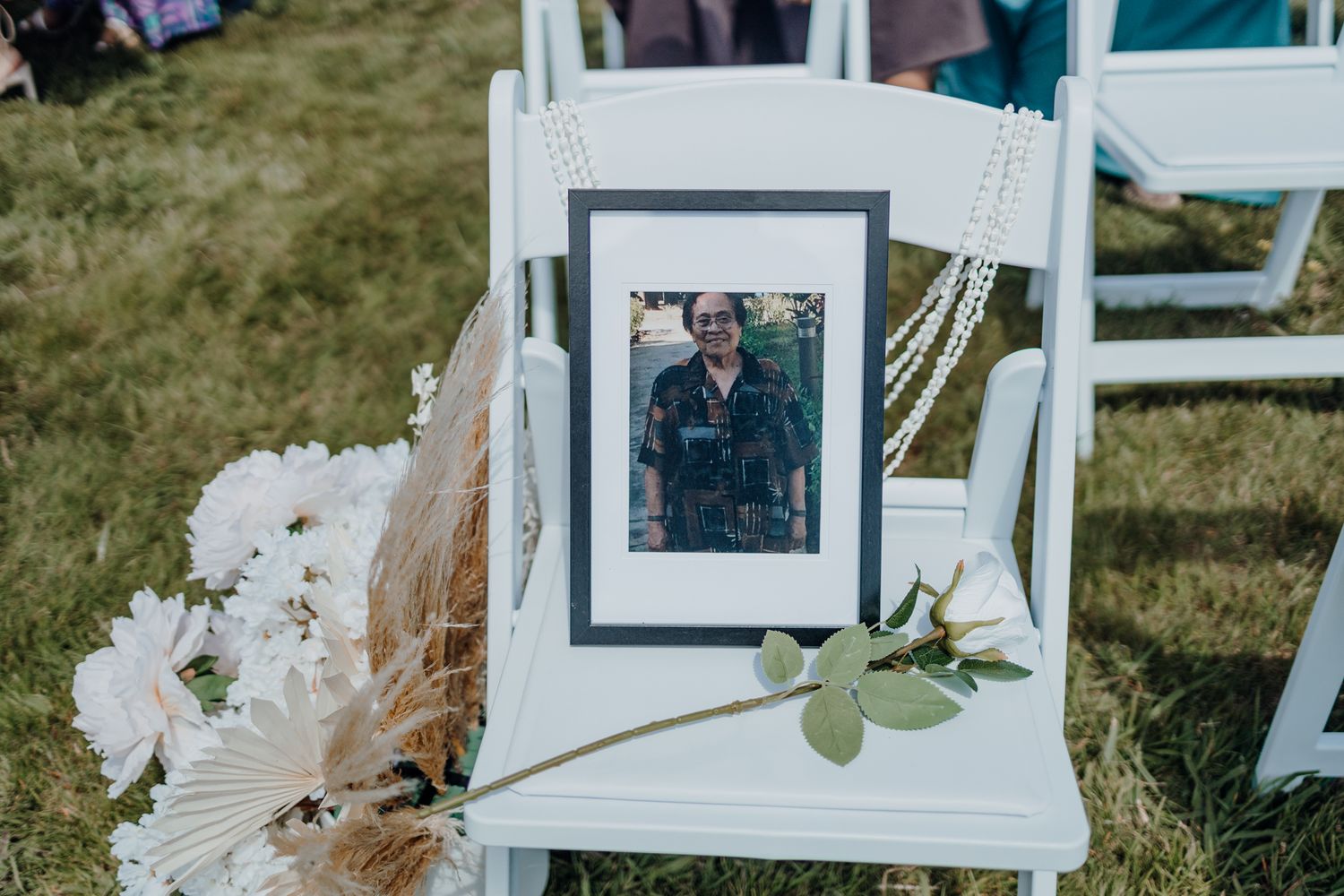 Framed memorial photo displayed on white ceremony chair with floral decorations and dried pampas grass.