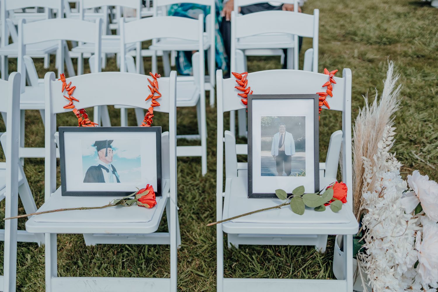 Memorial photographs displayed on white ceremony chairs decorated with red flowers and pampas grass.