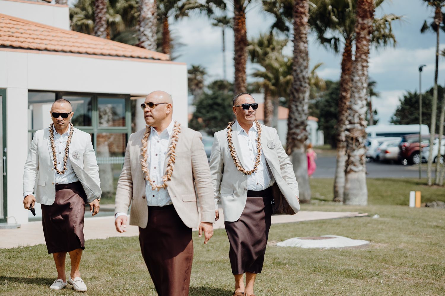 Wedding party members in matching light gray jackets walk along a palm-lined path.