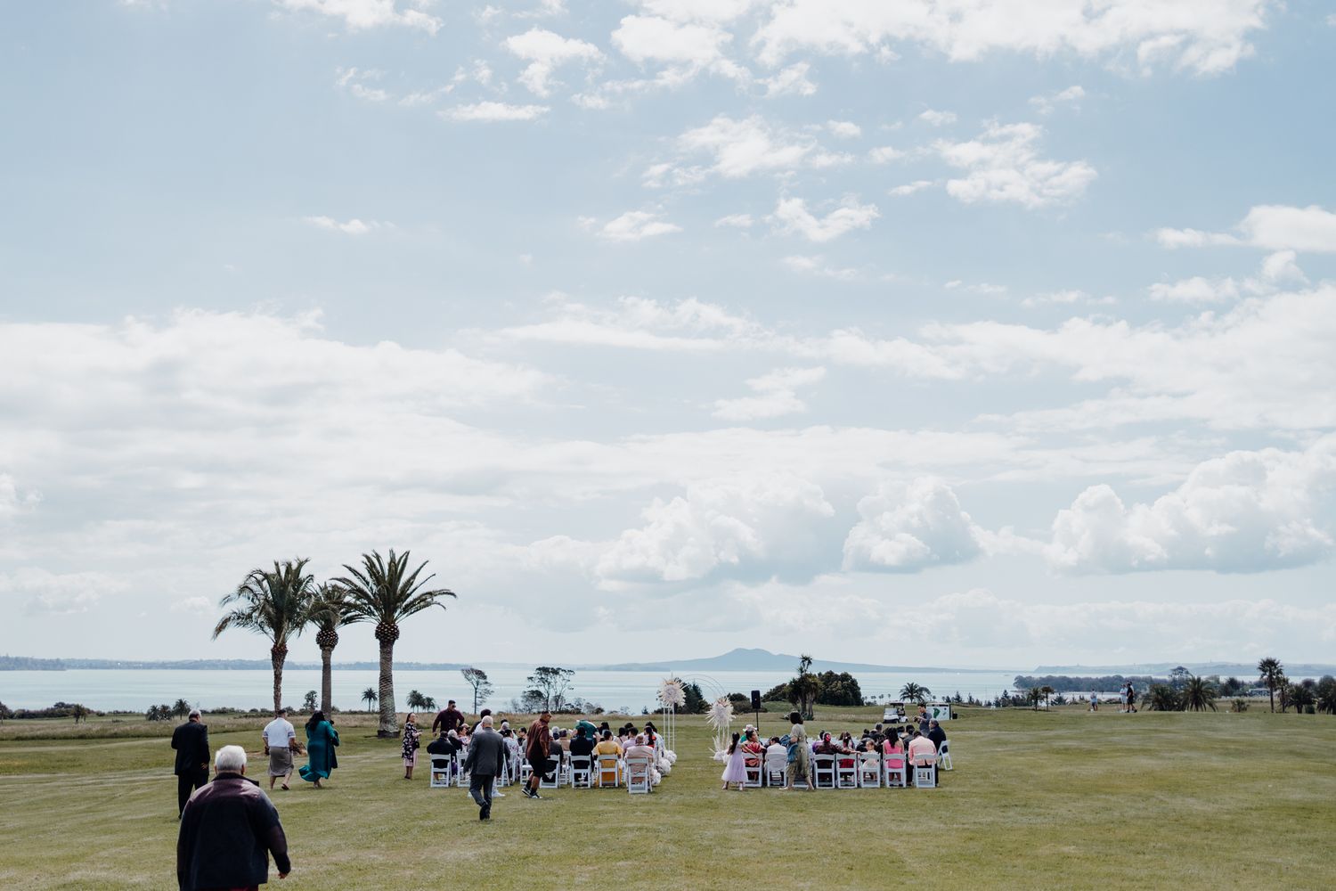 Wedding ceremony gathering on a coastal lawn with palm trees and ocean views in the background.