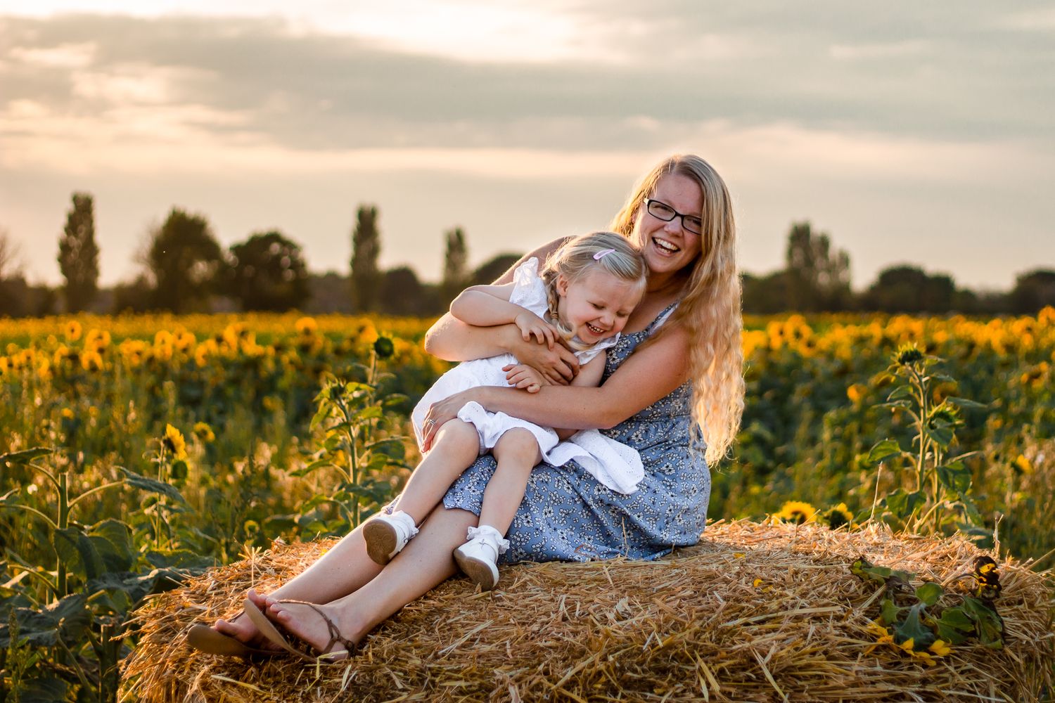 In a Sunflower field on Hayling Island, Hampshire a Mum and her two year old Daughter sit on a hay bale laughing.