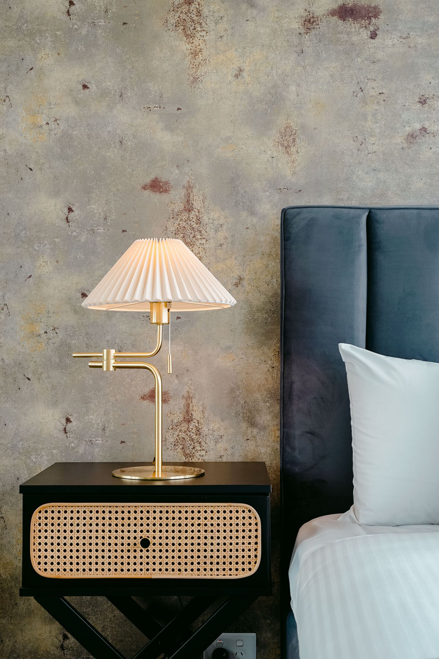 Modern bedside table with gold lamp and cane detailing against distressed concrete wall.