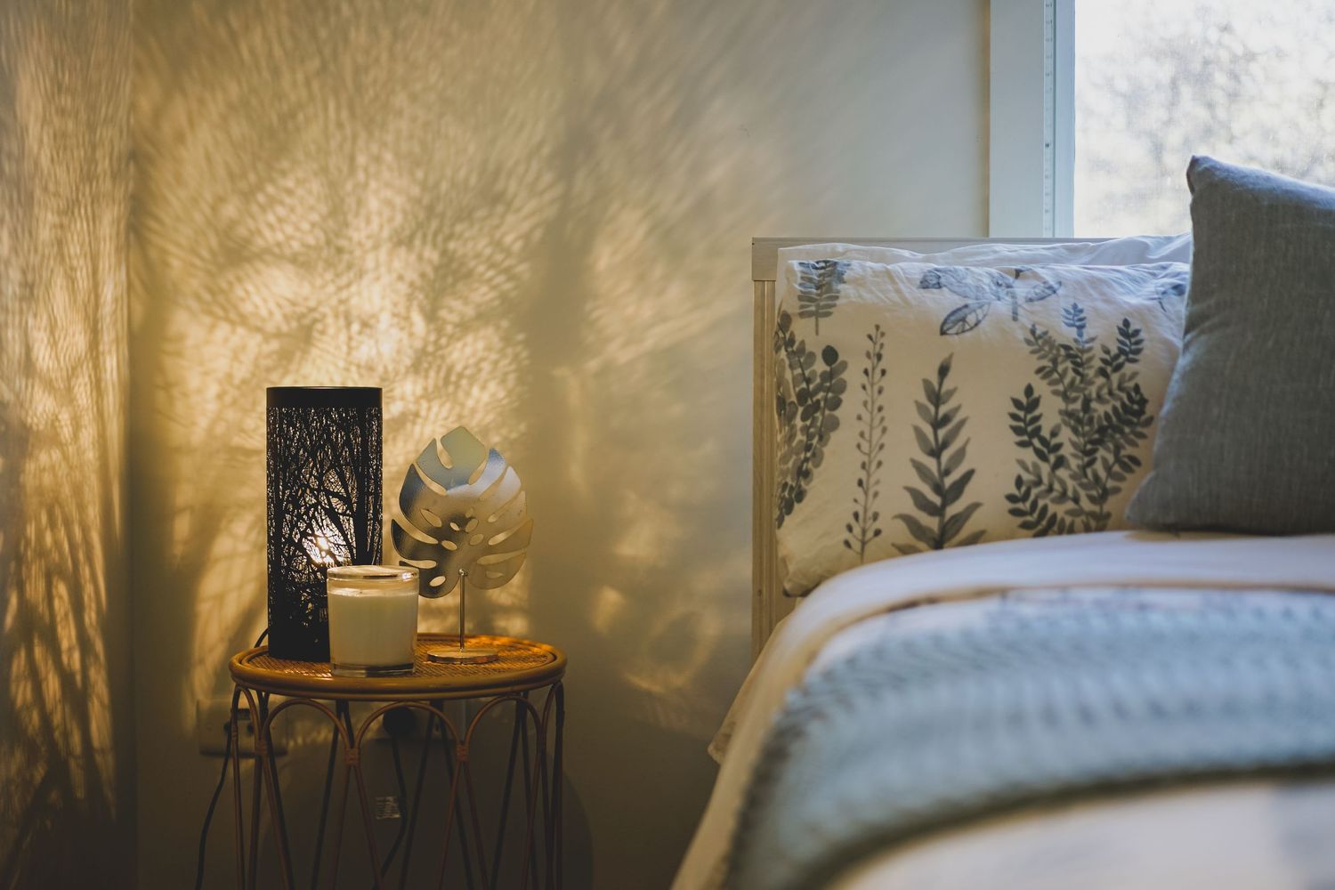 Warm bedside lamp casting botanical shadow patterns on textured wall beside cozy bedding.