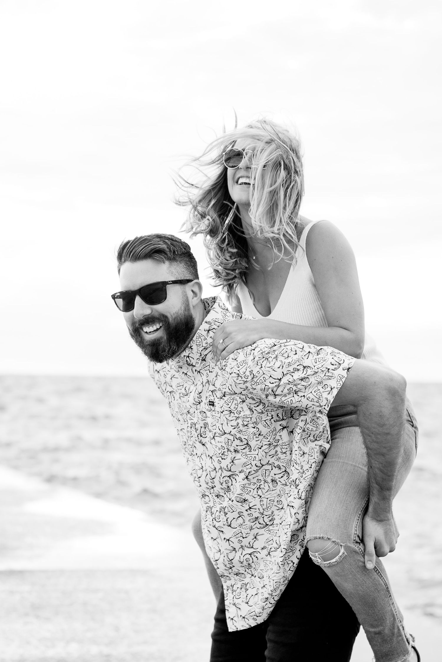 woman in white tank top and sunglasses piggyback riding on her dark haired fiance in sunglasses at North Ave Beach