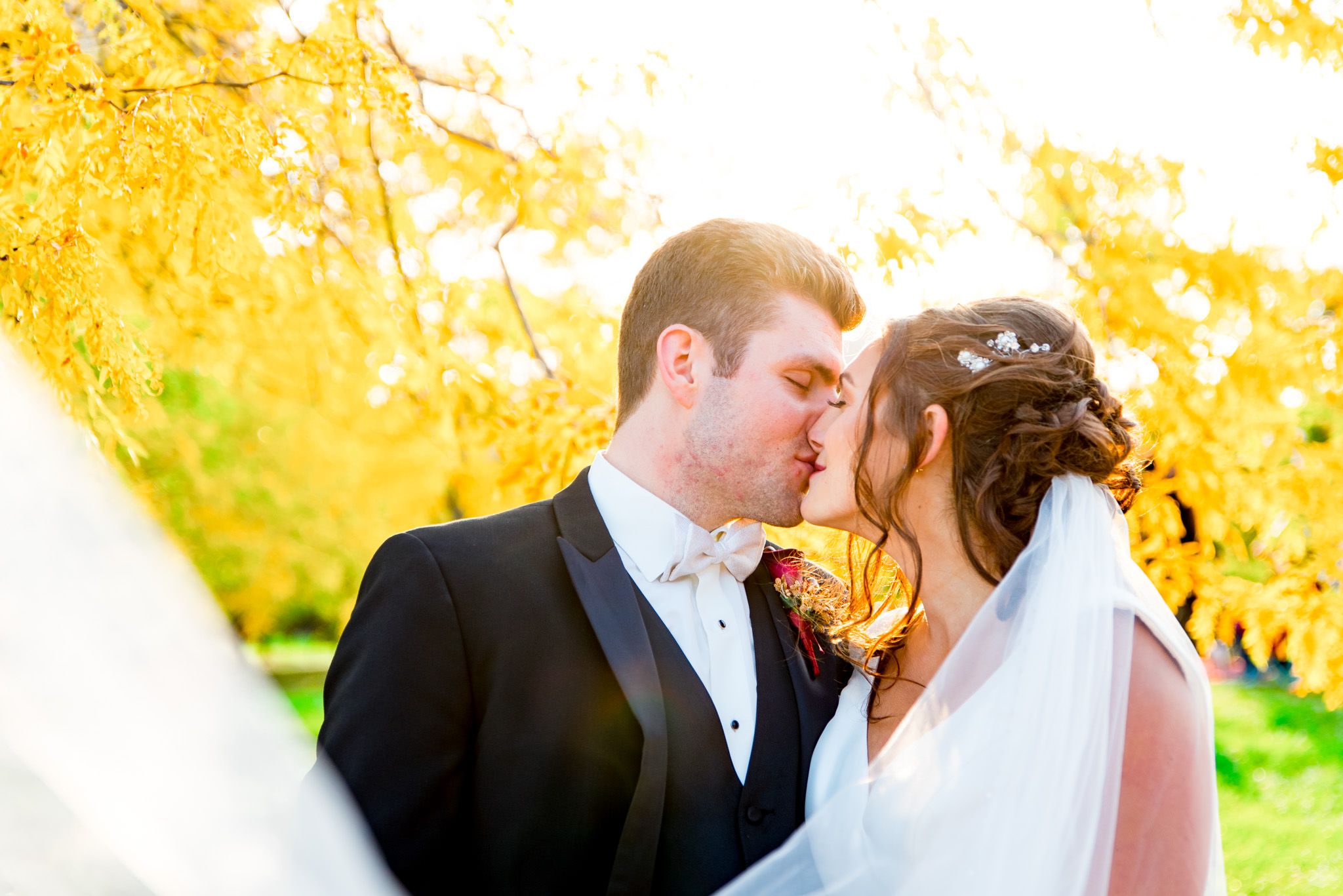 bride wears white wedding veil and kisses her husband in front of fall leaves in chicago park