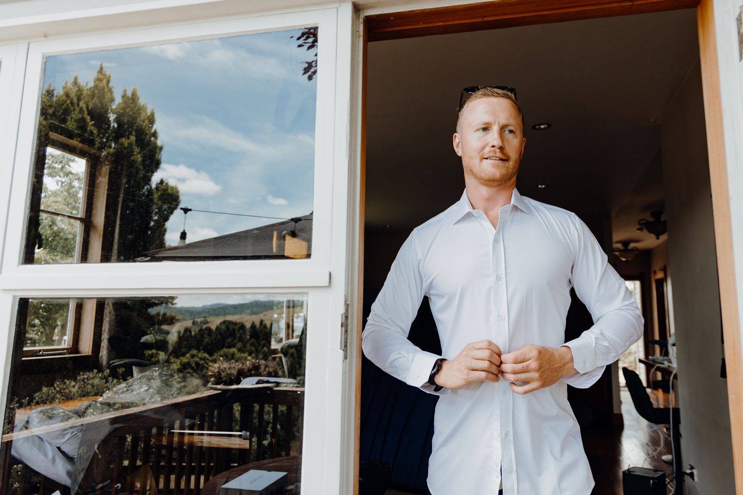 A person adjusts their light blue dress shirt while standing in a doorway with a window view.
