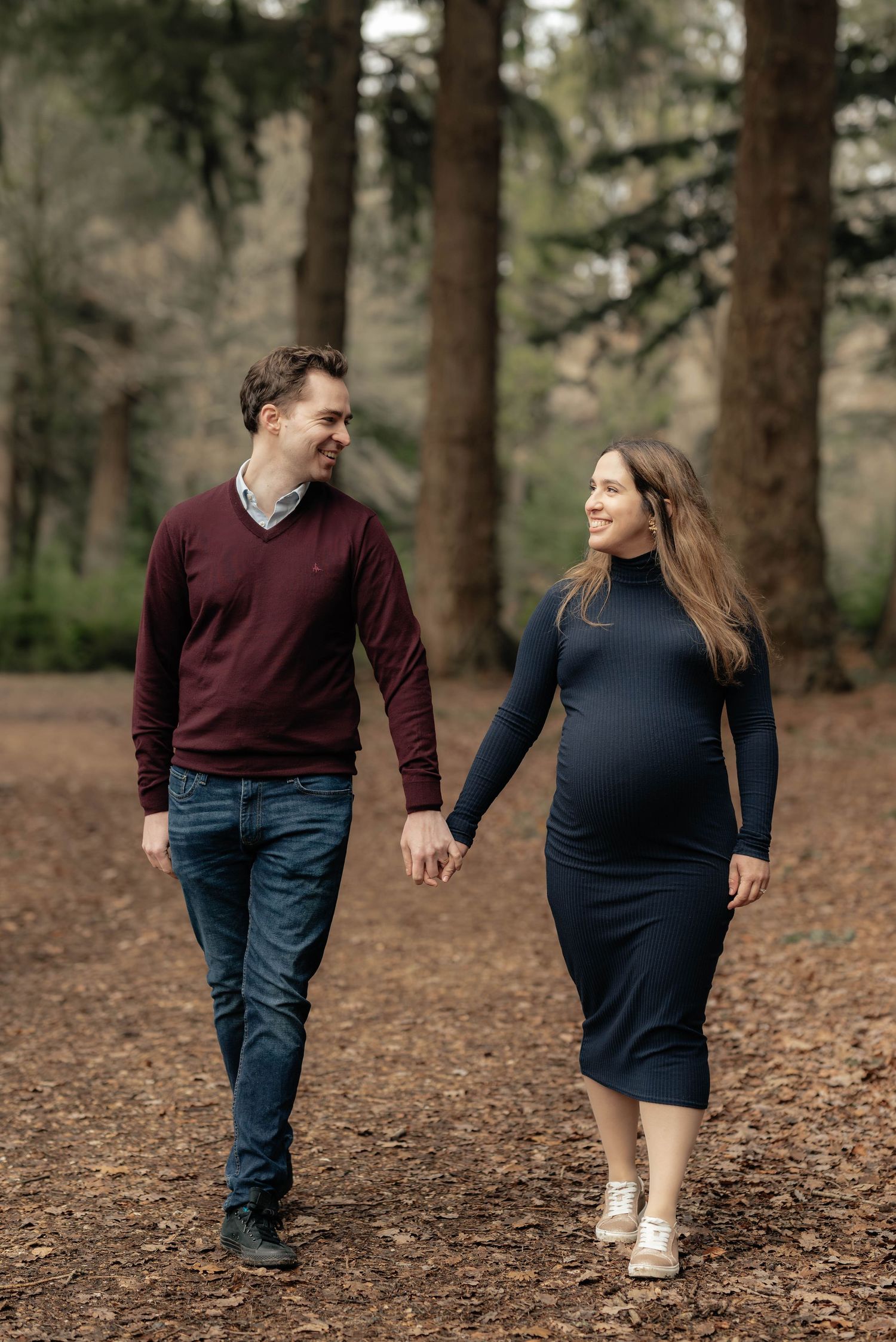 Couple walks hand in hand through forest setting with pine trees in background.