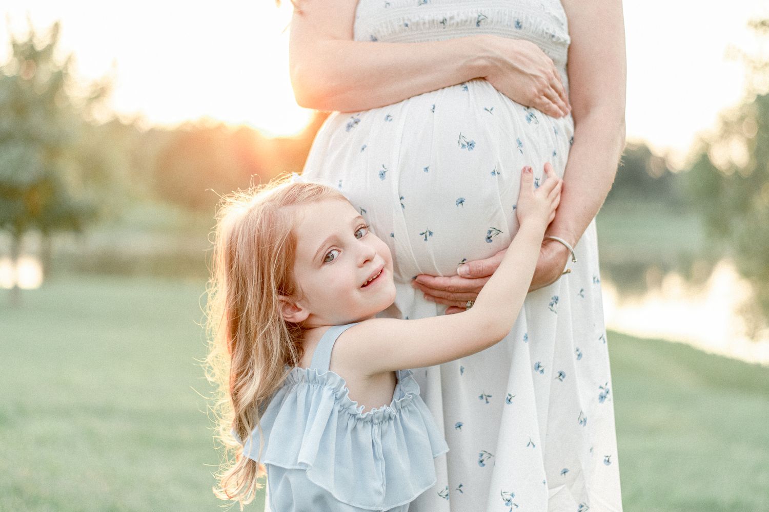 Little girl hugs her mother's pregnant stomach with golden hour light behind them