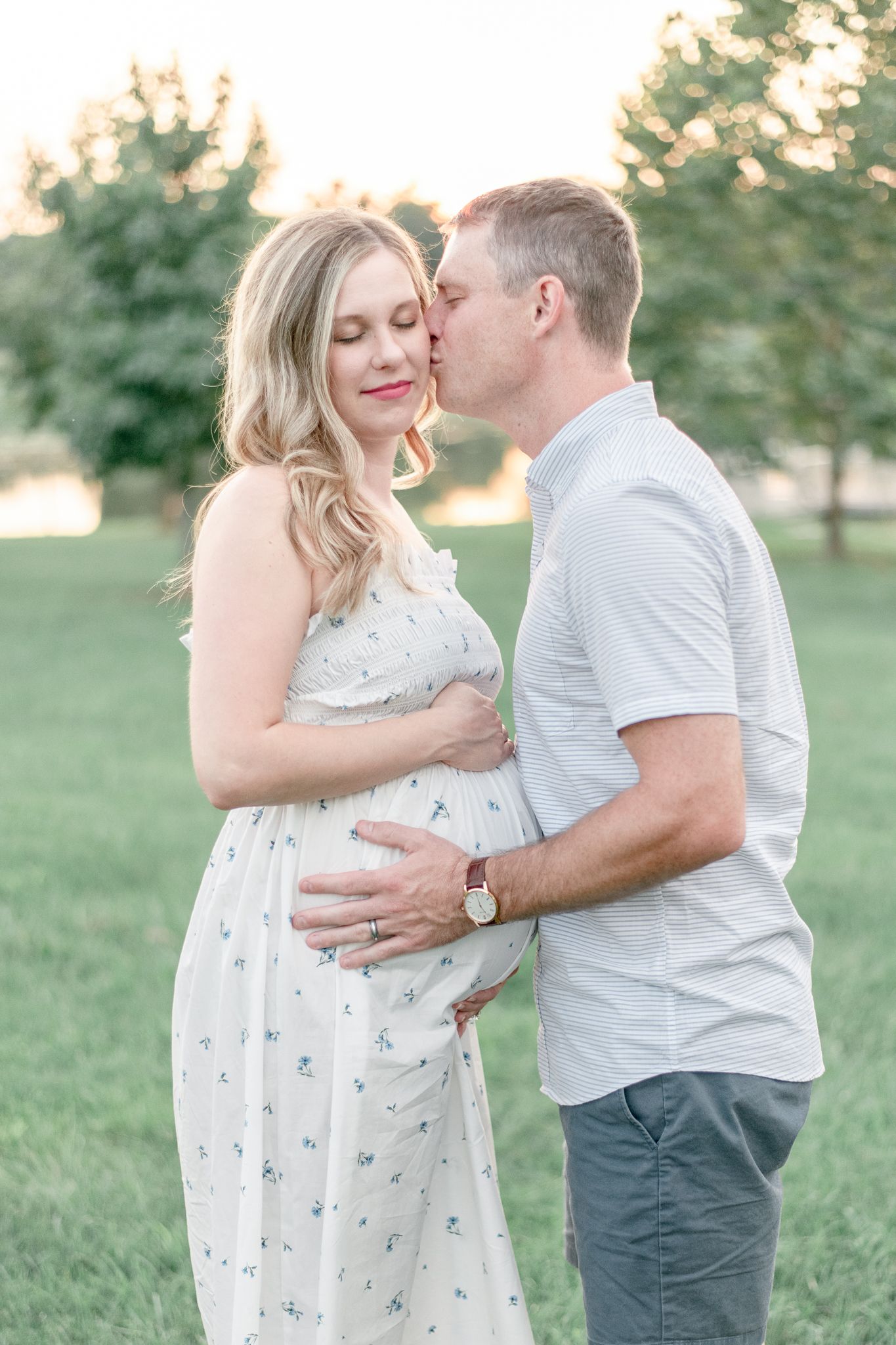 Man holds his wife's pregnant stomach and kisses her on the cheek at sunset