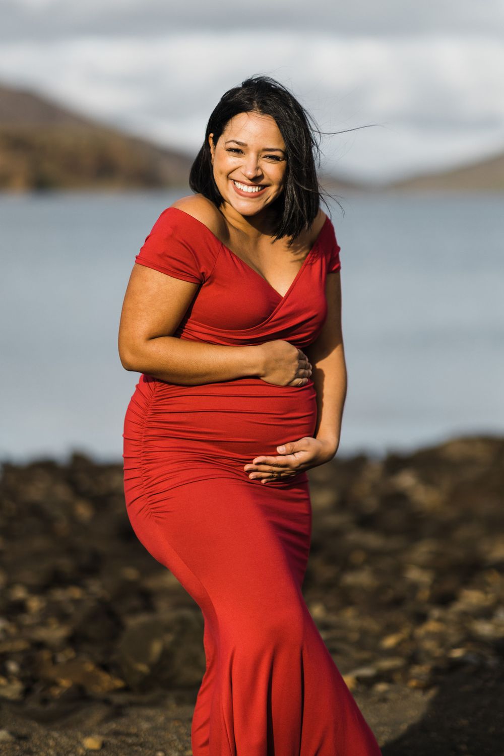 Maternity portrait in red dress against scenic mountain lake backdrop.