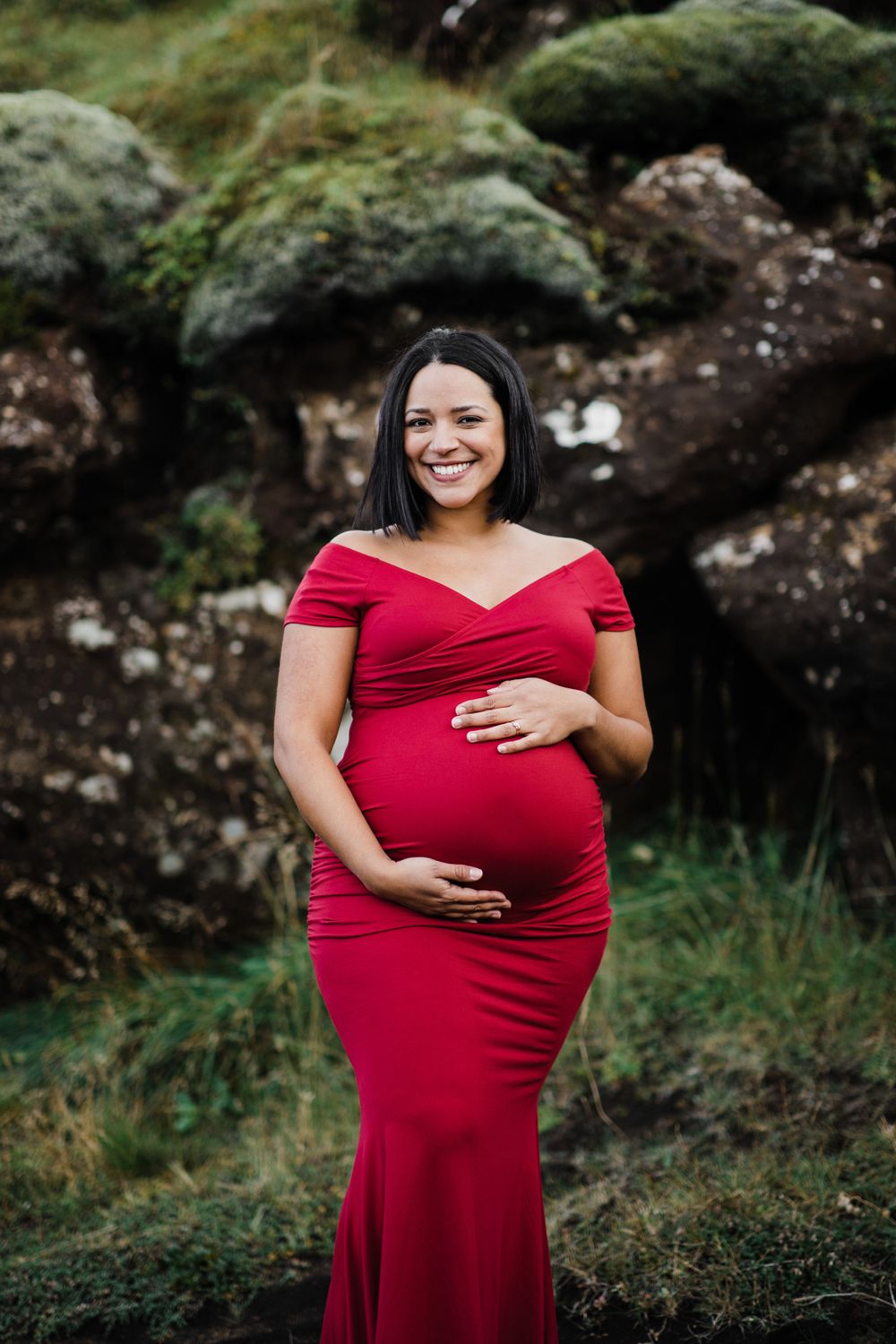Expectant mother in flowing red dress poses against mossy rocks during maternity photoshoot.