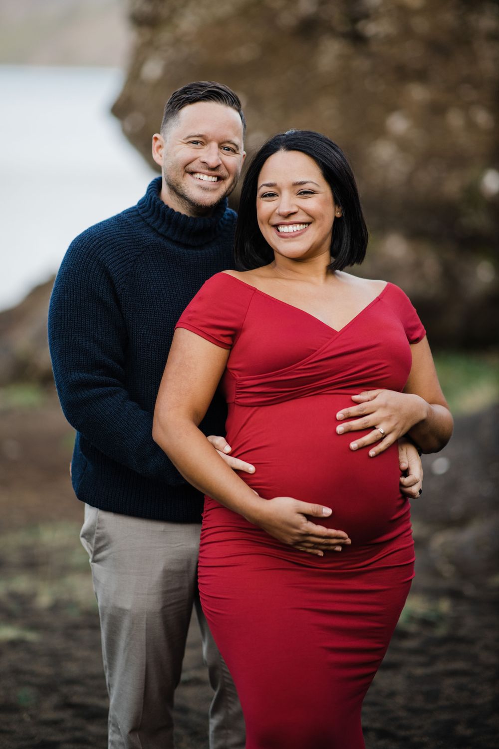 Happy couple embracing during maternity photo session, wearing navy sweater and red dress.