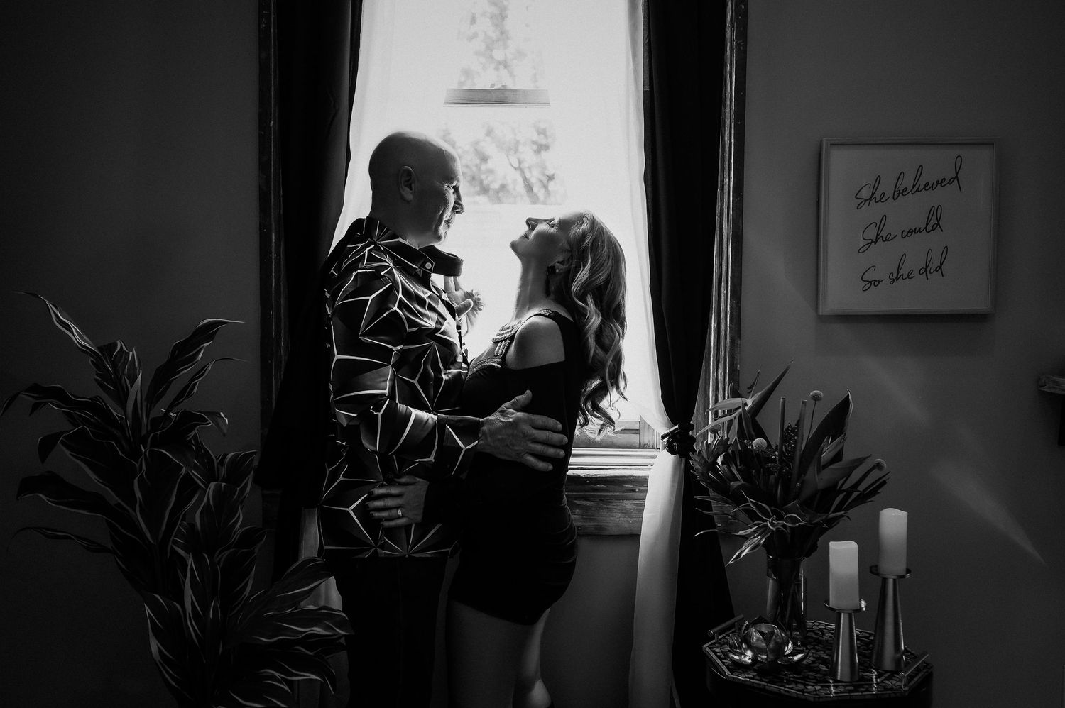Intimate black and white silhouette of a couple by a window with plants and candles.