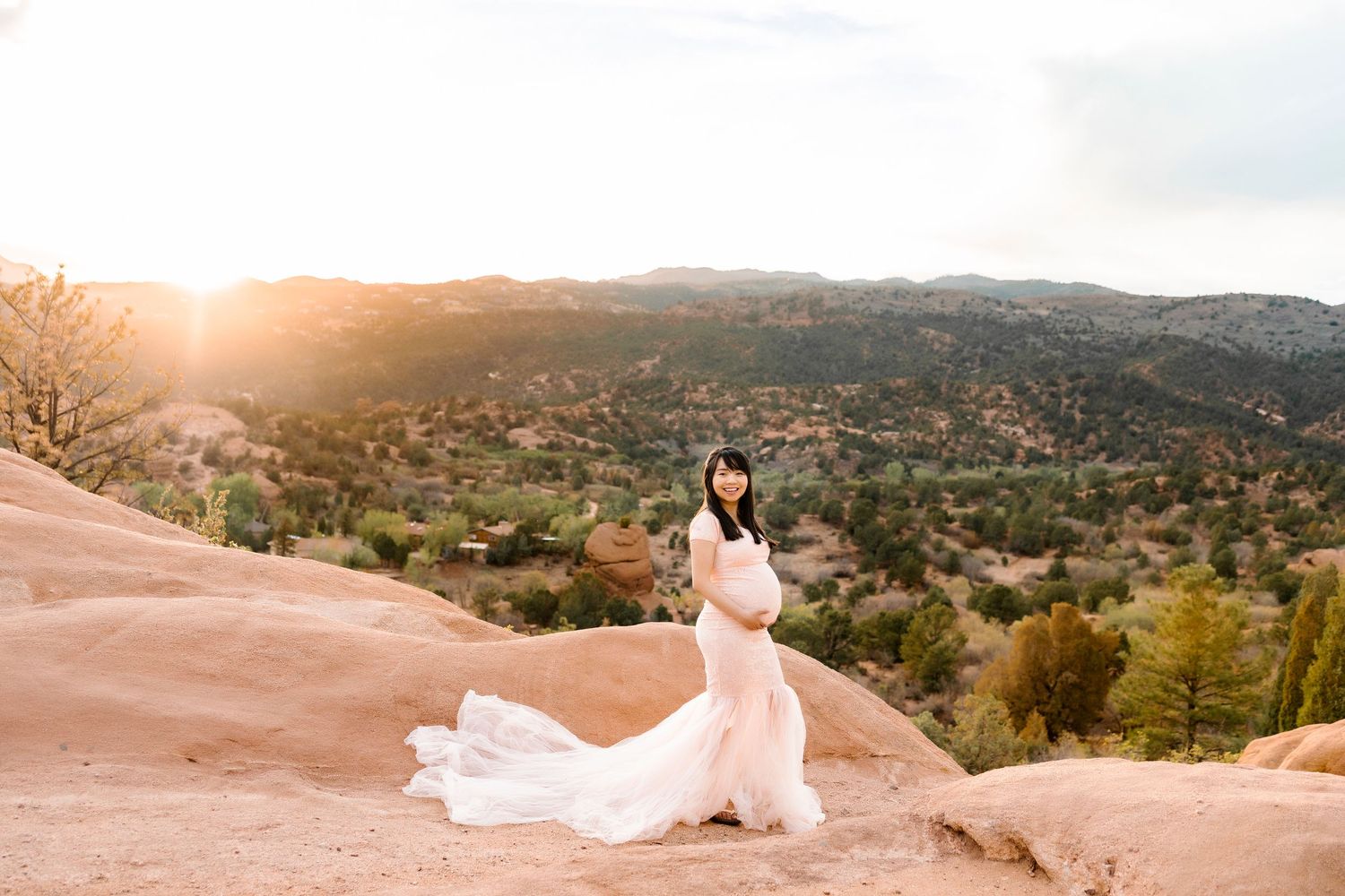 Pregnant woman in long pink dress on pink mountain, with green landscape behind, sunsetting