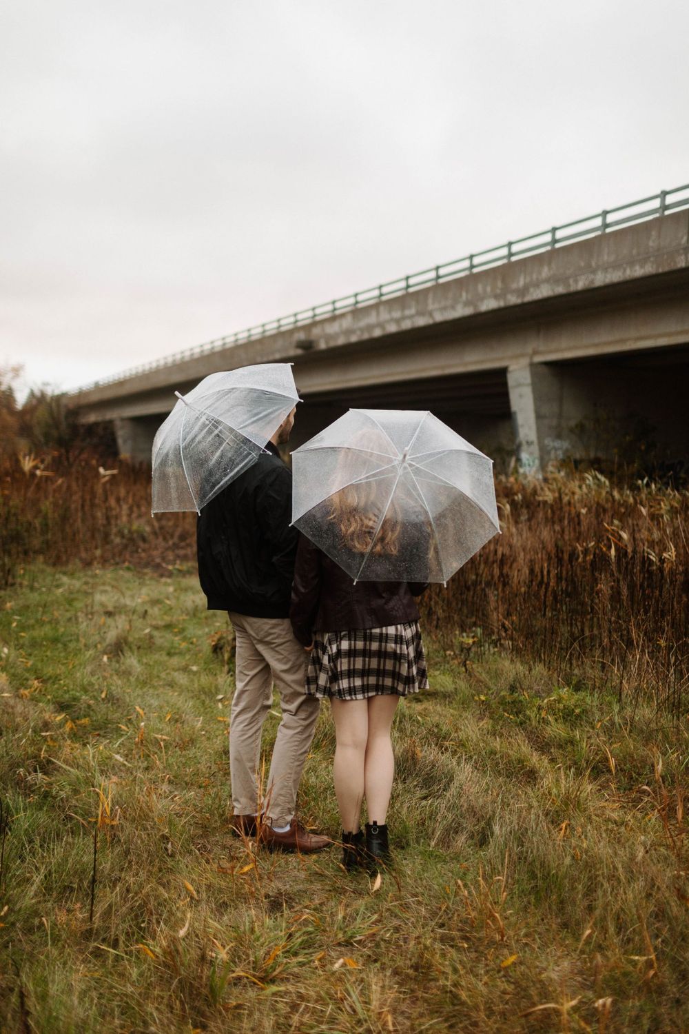 Couple holding clear umbrellas stands together in rainy field near concrete bridge in Kitchener, Ontario