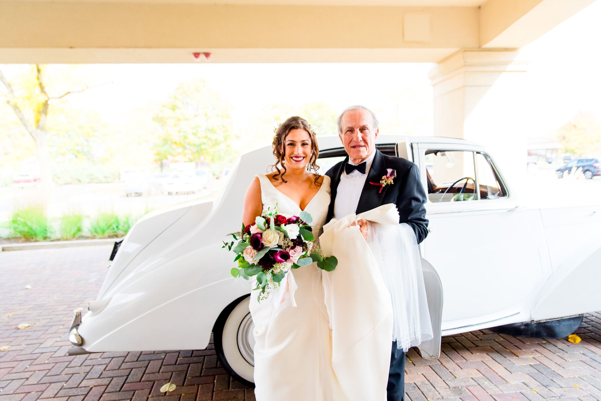 bride with burgundy and white flowers poses with her father in front of a bentley