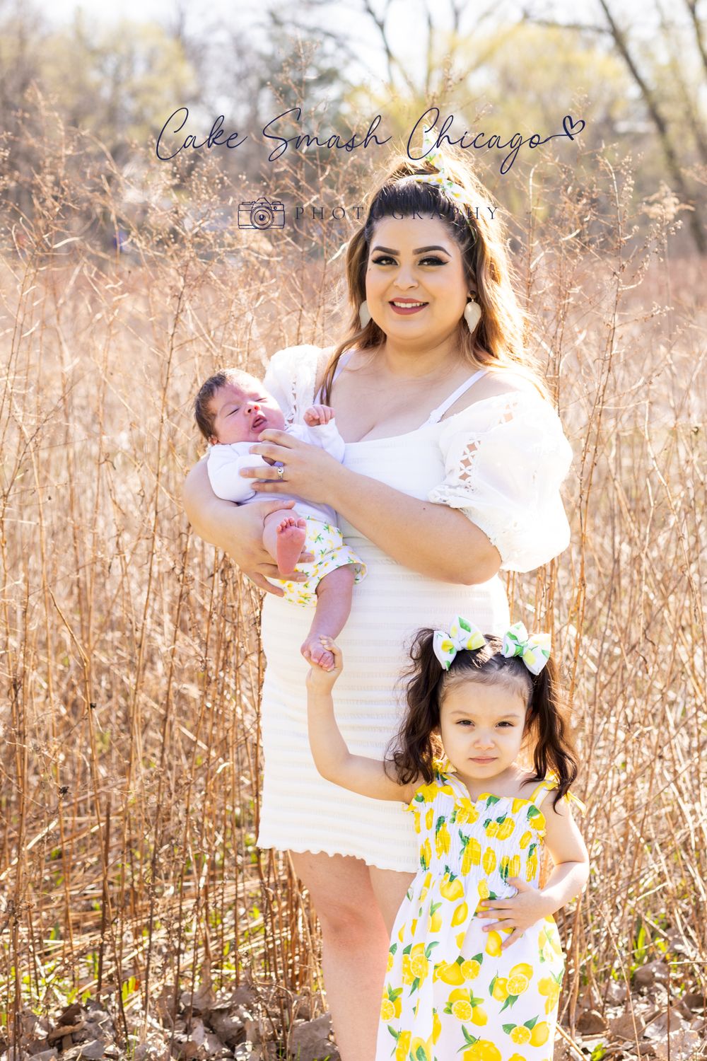 A spring outdoor family portrait featuring a woman in white dress with two children amid tall grass.