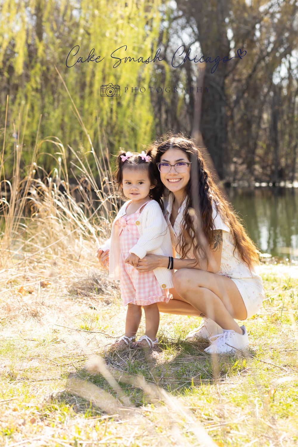 Mother and daughter pose together by lake with willow trees in background.