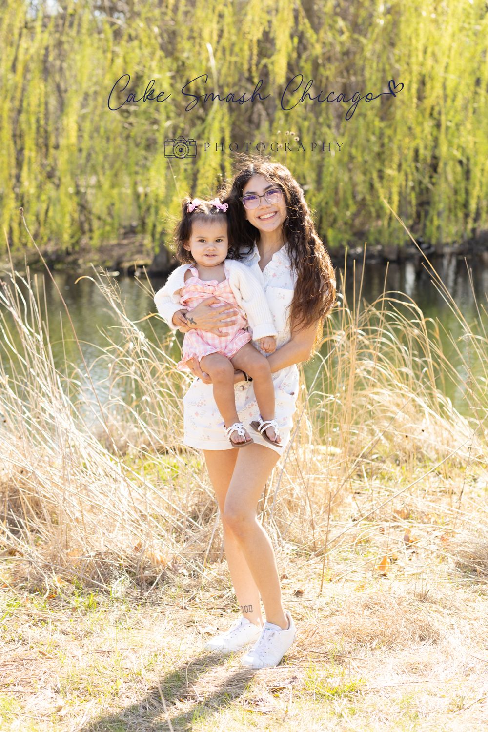 Sunlit portrait of mother and child standing among tall grass near water.