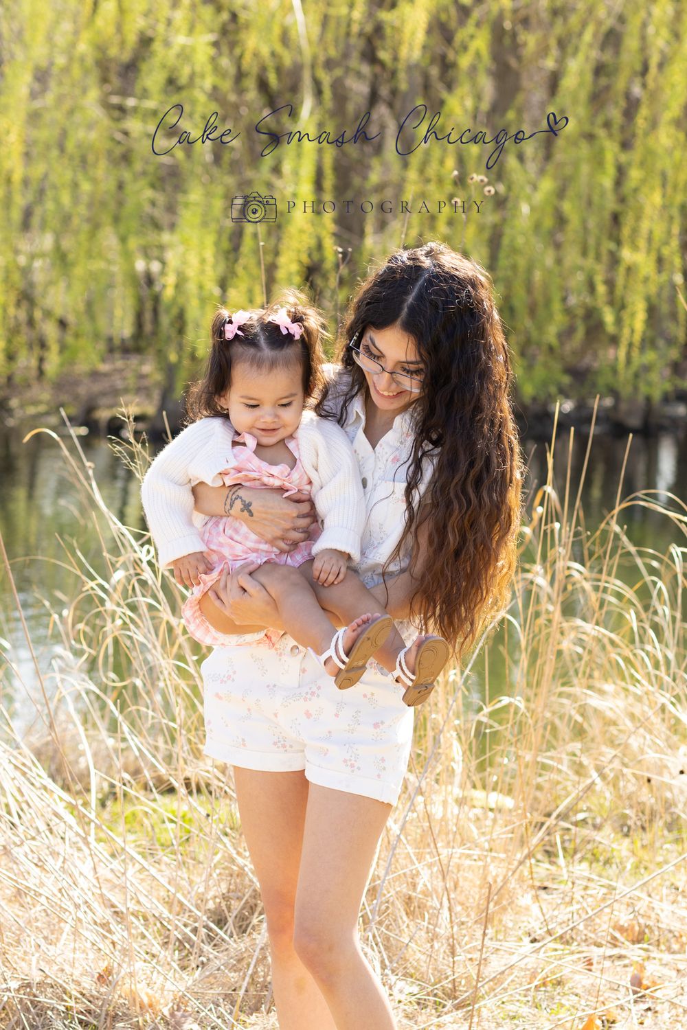 A parent and young child share a tender moment together among tall grass and weeping willows on a sunny day.