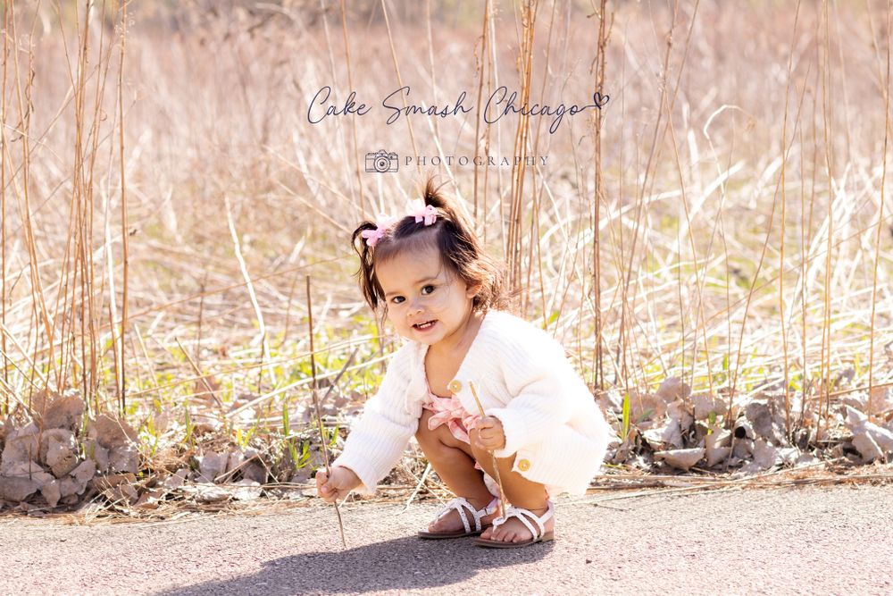 A toddler in a white cardigan and pink dress crouches curiously on a pathway outdoors.