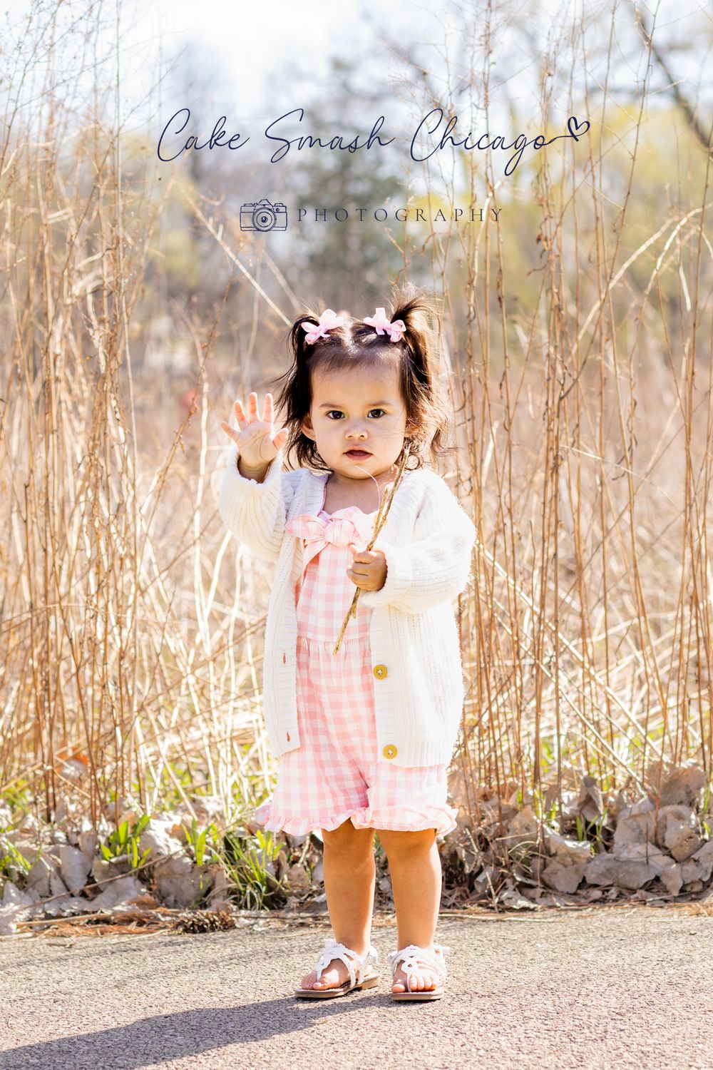 A small child in a white cardigan and pink dress stands amidst dried grass and natural scenery.
