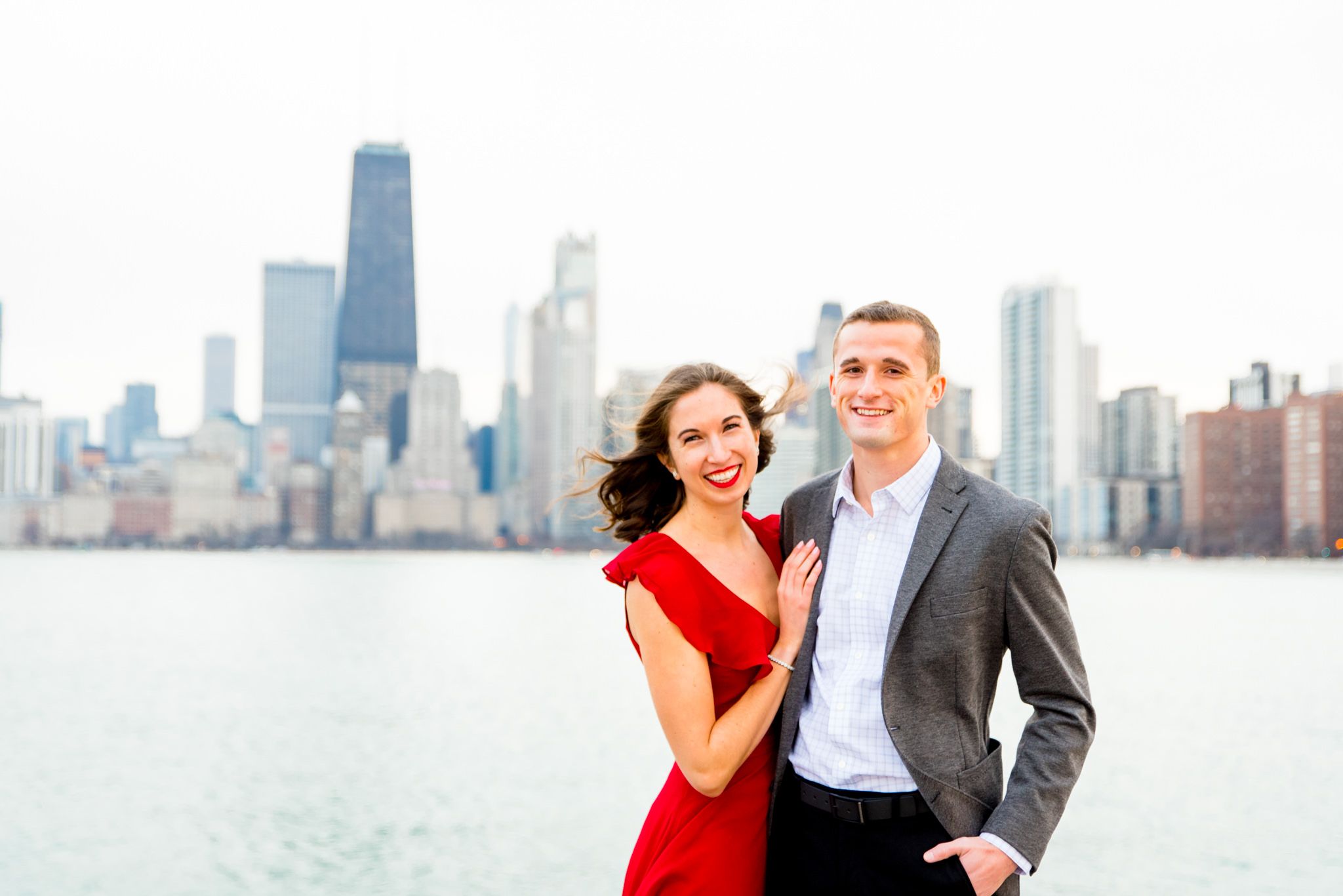 brunette in red dress and fiance in grey suit smiling in front of downtown Chicago for engagement pictures in Chicago