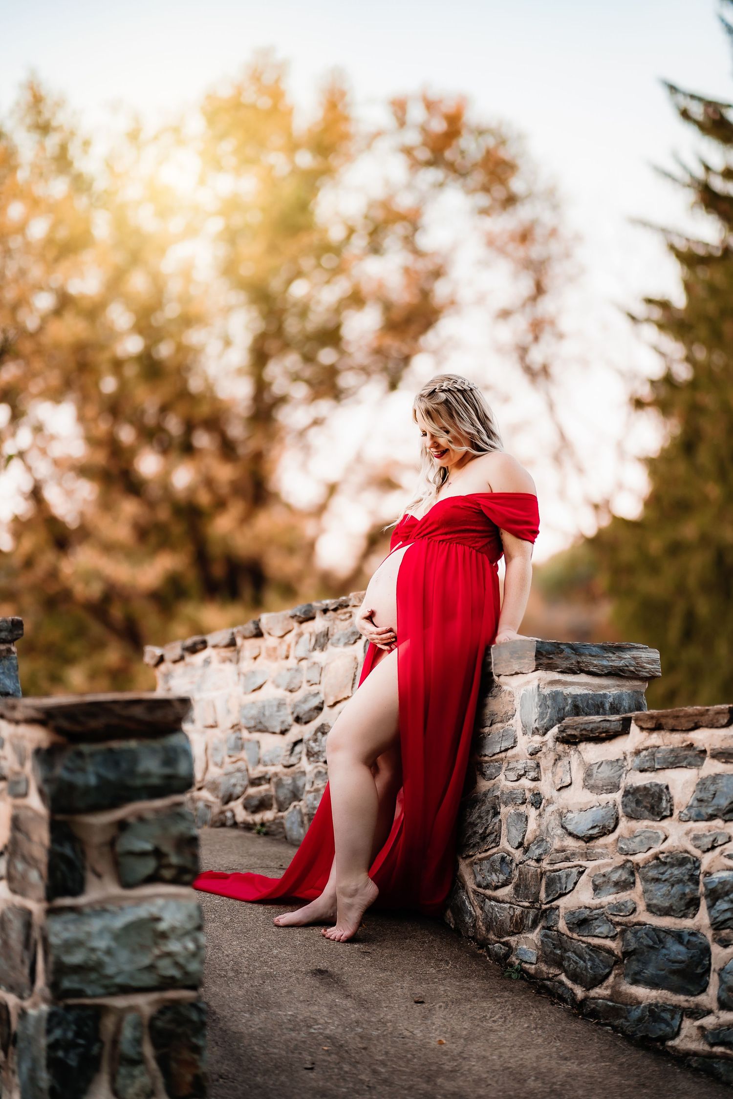 Maternity portrait of woman in flowing red dress against stone wall during golden hour.