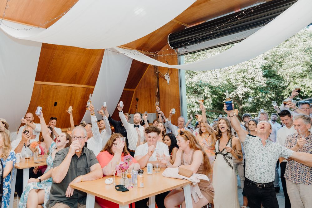 Wedding reception guests cheer with raised arms in modern venue with wood panels.