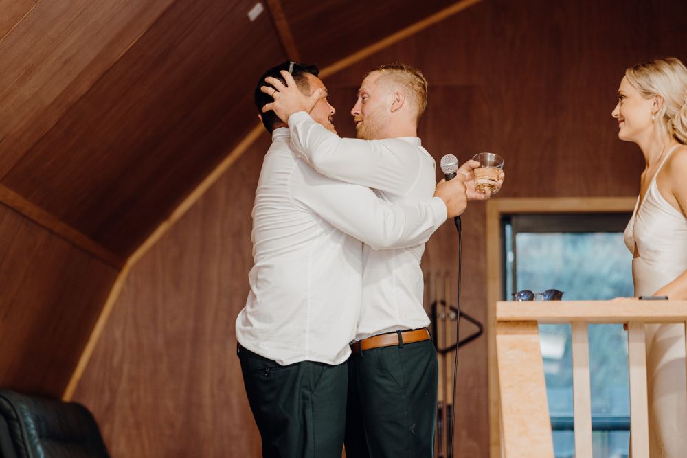 Two people embrace while another person watches from the side in a wooden interior.