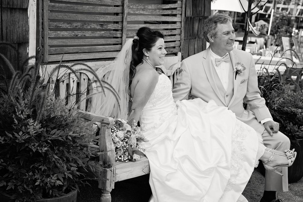 Black and white photo of couple sitting together on rustic bench in wedding attire.