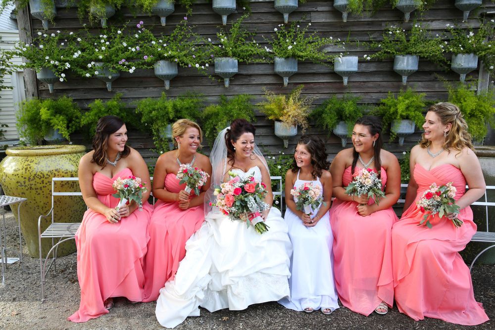 Wedding party in coral dresses seated on garden bench with floral wall backdrop.