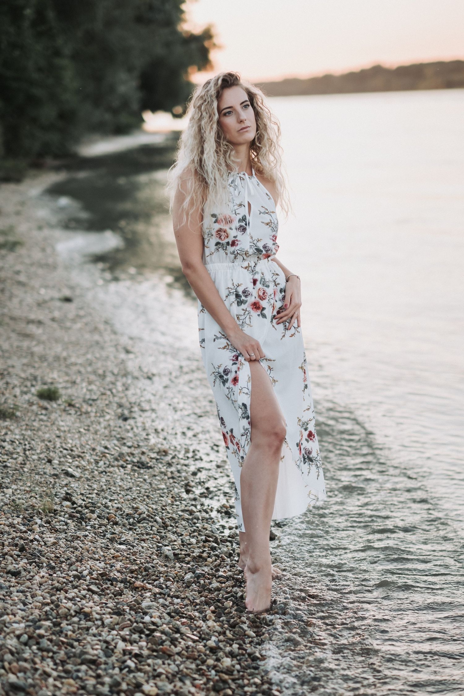 Woman in flowing floral dress poses elegantly on rocky shoreline at sunset.