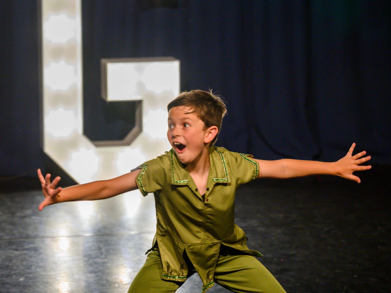 A performer in a green costume strikes an energetic pose with arms outstretched on a dark stage.