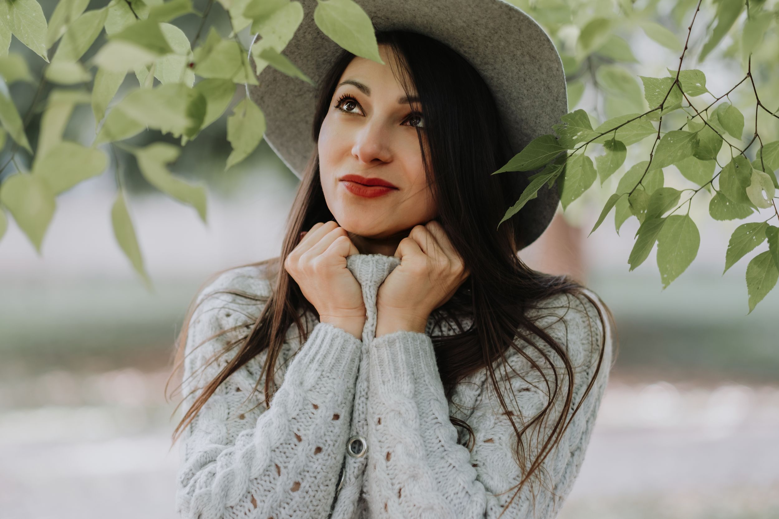 Outdoor portrait under green leaves wearing a gray sweater and wide brim hat.