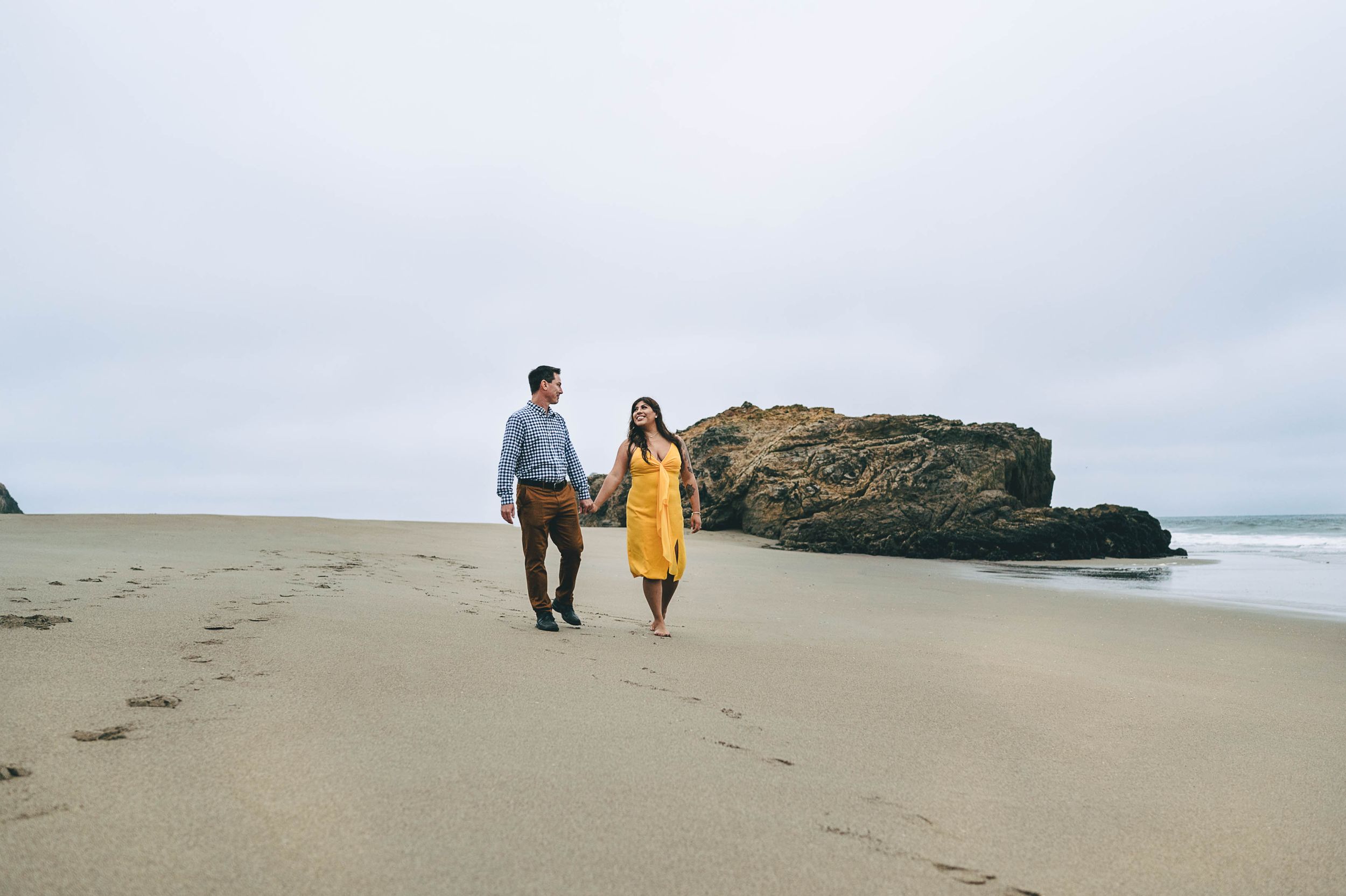 Ryan and Nataly in her yellow dress walking along the beach at sunrise under moody sky.