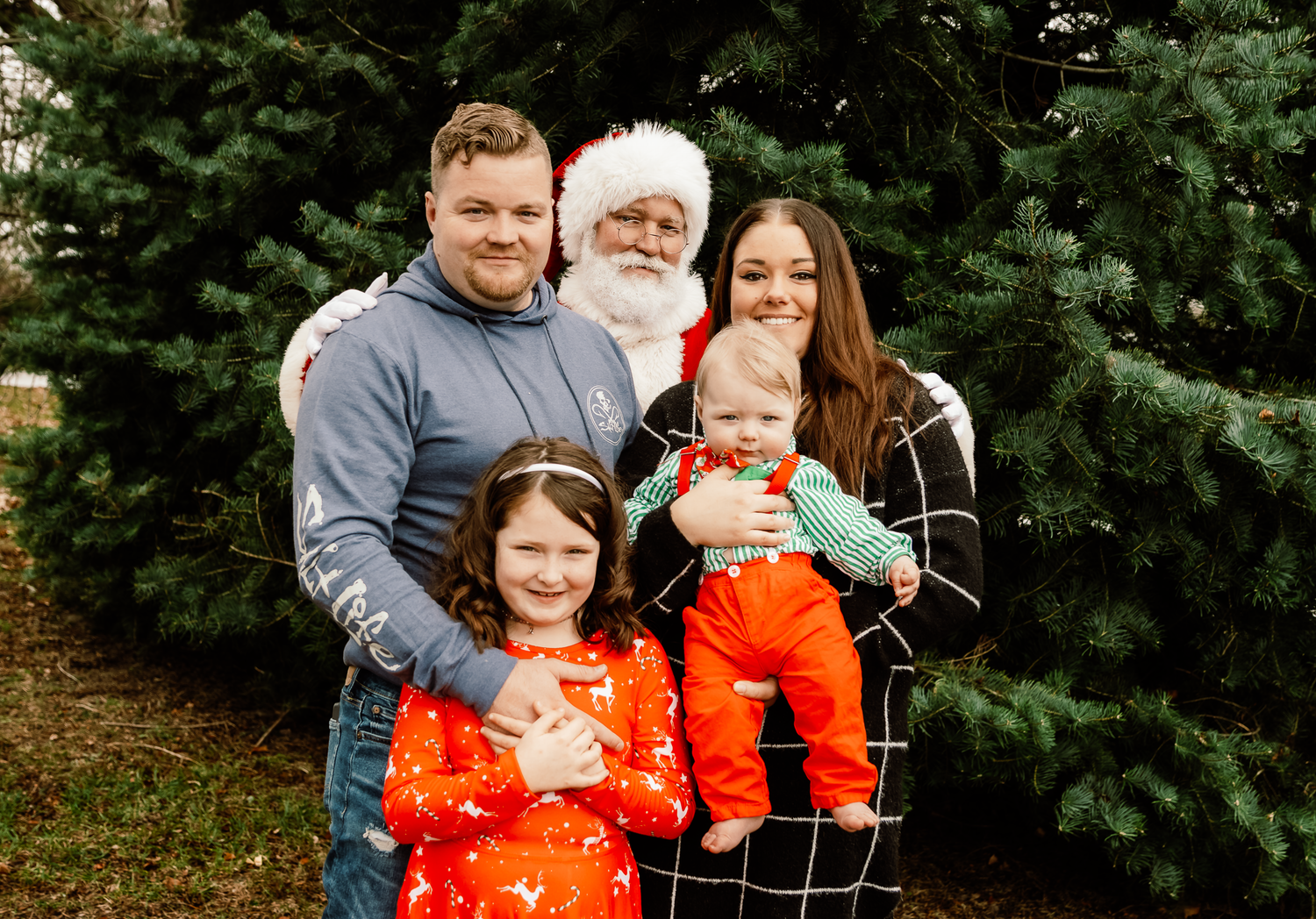 Holiday family portrait with Santa Claus and evergreen trees in the background.