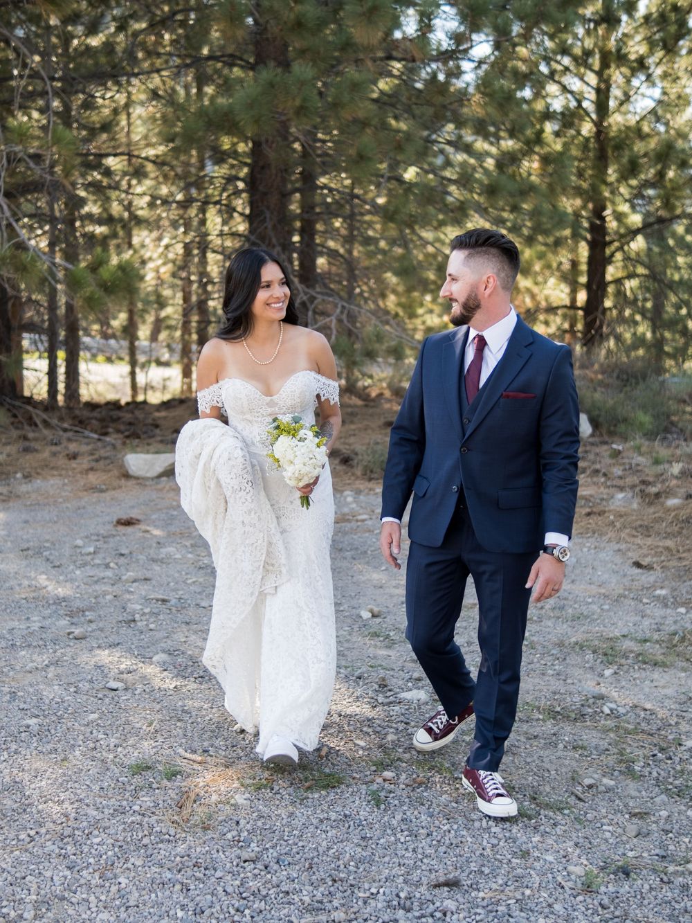 A couple walks along a mountain path with pine trees, bride in white dress and groom in navy suit with casual sneakers.