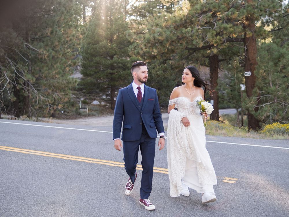A couple walking together on a tree-lined road, with the groom in a navy suit and bride in a flowing white gown.