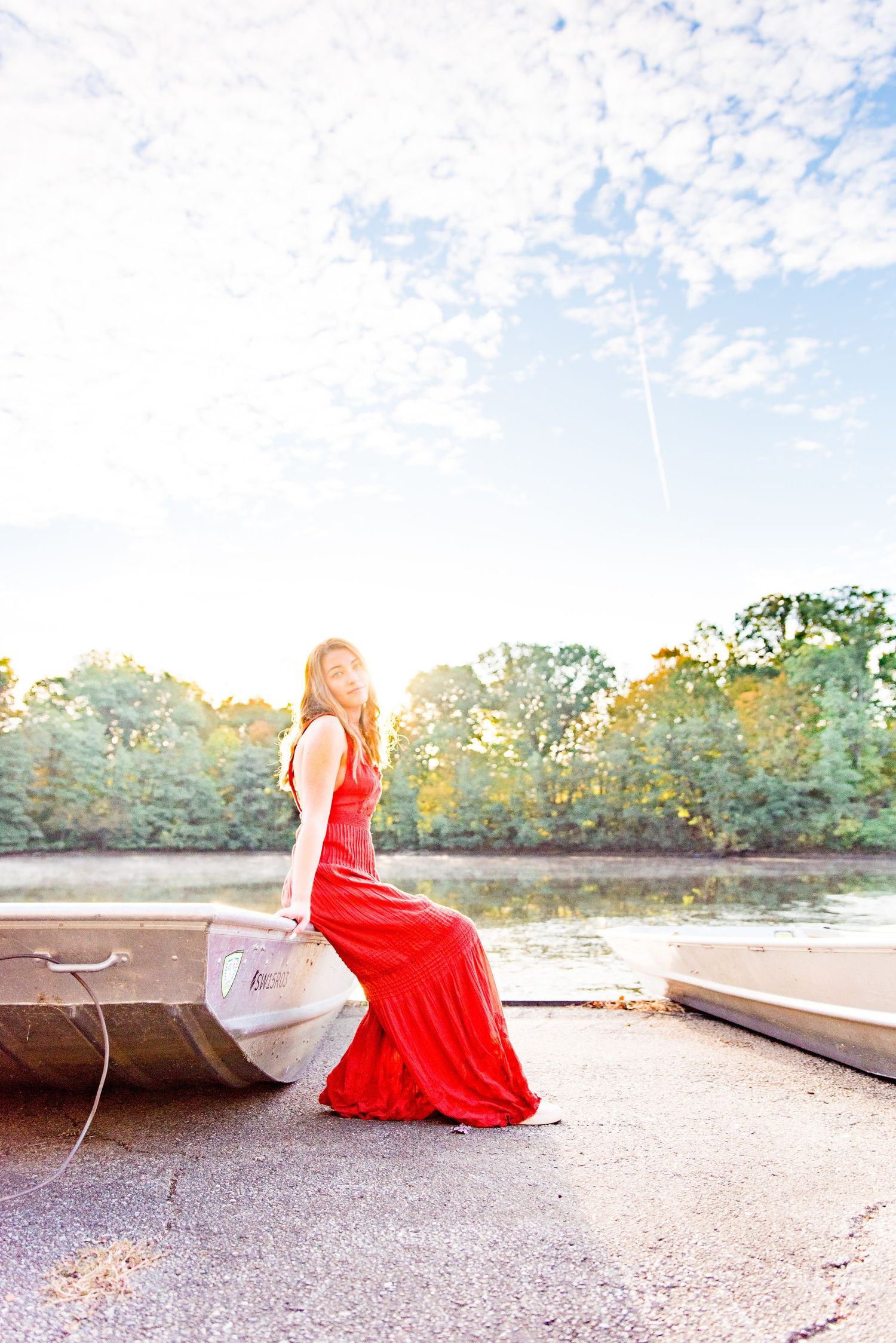 young woman in red jumpsuit sits on edge of boat in front of a pond under a blue cloudy sky at Sharon Woods Park