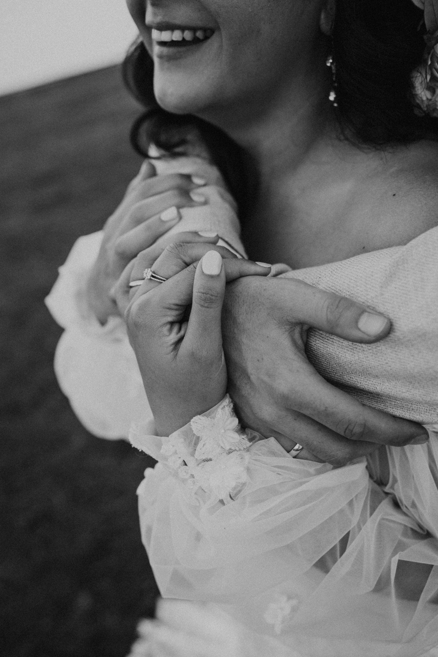 Black and white close-up of hands with wedding rings and lace dress details.