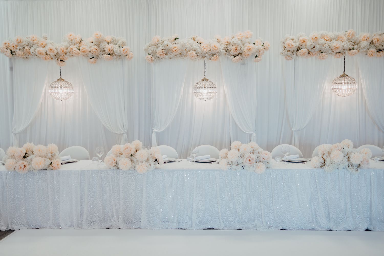 Elegant white wedding reception setup with hanging pendant lights and white floral arrangements.