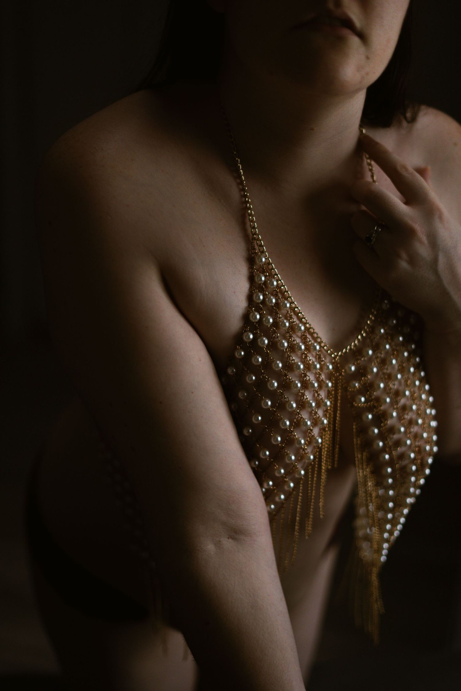 moody photo of a dark haired person wearing a gold and pearl body chain looking towards the camera