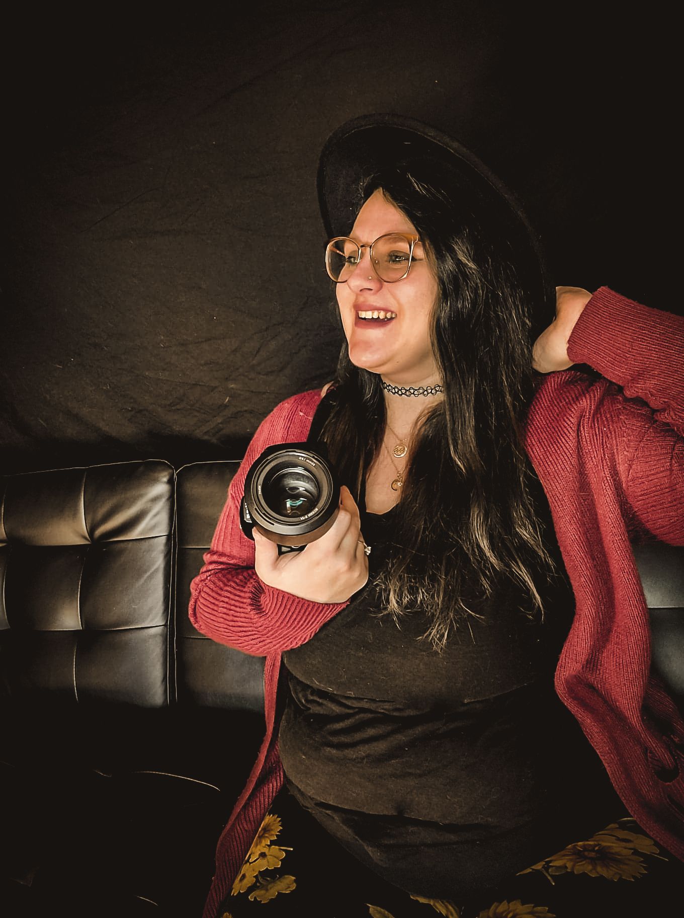 Photographer in red cardigan and black hat laughing while holding camera on leather couch.